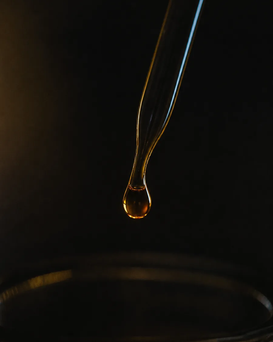 A single amber droplet hanging from the tip of a glass pipette above a dark dish.
